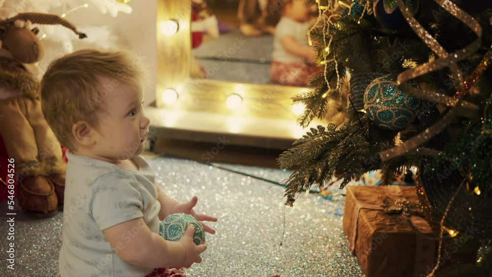 Cute little baby boy sit under a fir-tree with wrapped gifts and bright lights holding a xmas ball in hands. Sweet child hanging christmas decorations at home. New year winter holiday, family concept.