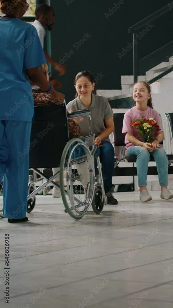 Vertical video: Mother and child visiting old woman in wheelchair at medical facility, talking to senior patient with disability. Little girl bringing flowers to grandmother in hospital reception