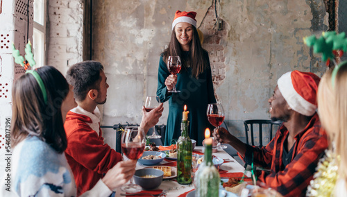 Young woman stands with a glass and says a toast while friends sit at the festive table.