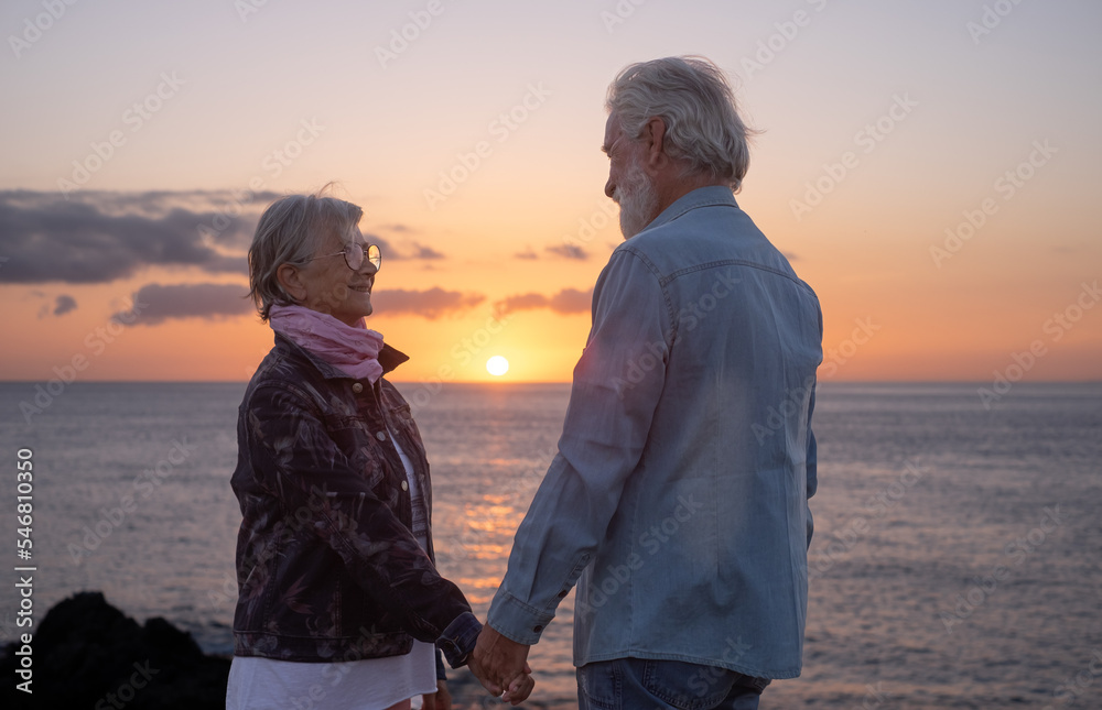 Fototapeta premium Lovely senior couple standing on the rocks at sea hand in hand enjoying nature and freedom at sunset light. Relaxed lifestyle for a caucasian couple of retirees
