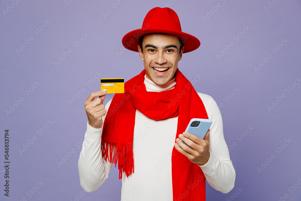 Young middle eastern man wear white turtleneck red hat scarf using mobile cell phone hold credit bank card do online shopping order delivery book tour isolated on plain pastel light purple background. Young middle eastern man wear white turtleneck red hat scarf using mobile cell phone hold credit bank card do online shopping order delivery book tour isolated on plain pastel light purple background.