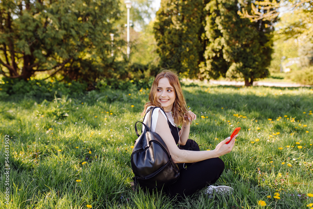 Photo of a positive cheerful teenage girl spends time in the park and using mobile phone.