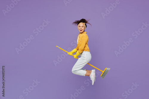 Fototapeta Naklejka Na Ścianę i Meble -  Full body side view smiling young housekeeper woman wear yellow shirt tidy up jump high sit on broom sweep floor look camera isolated on plain pastel light purple background studio. Housework concept.