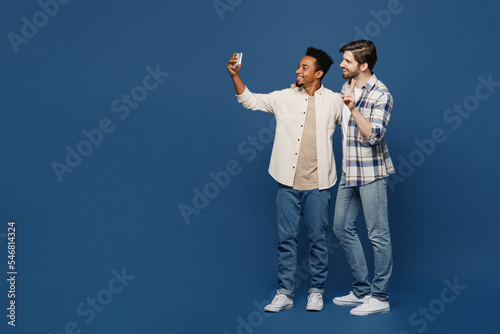 Full body young two friends happy men 20s wear white casual shirts together doing selfie shot on mobile cell phone show v-sign isolated plain dark royal navy blue background. People lifestyle concept.