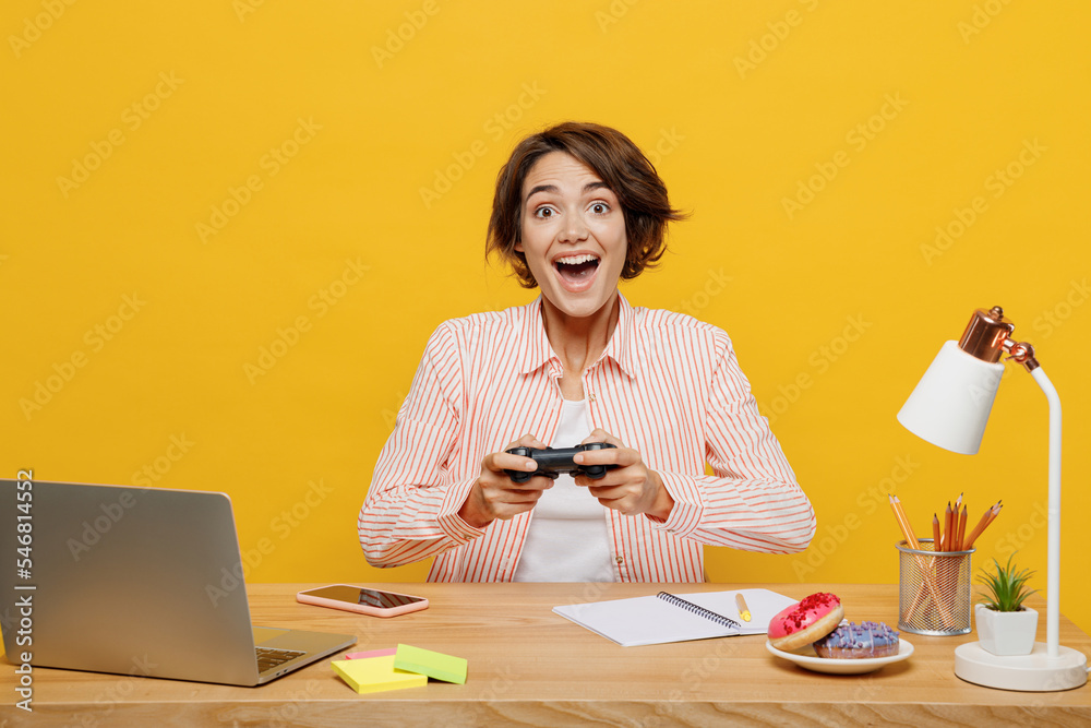 Young fun employee business woman wear shirt sit work at office desk with pc laptop hold in hand play pc game with joystick console in vr headset pc gadget isolated on plain yellow color background. Young fun employee business woman wear shirt sit work at office desk with pc laptop hold in hand play pc game with joystick console in vr headset pc gadget isolated on plain yellow color background.