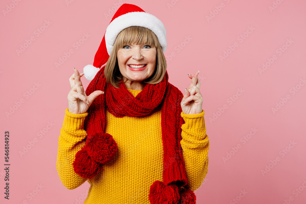 Merry elderly woman 50s year old in yellow knitted sweater red scarf Santa hat posing wait special moment keep fingers crossed isolated on plain pink background Happy New Year Christmas 2023 concept