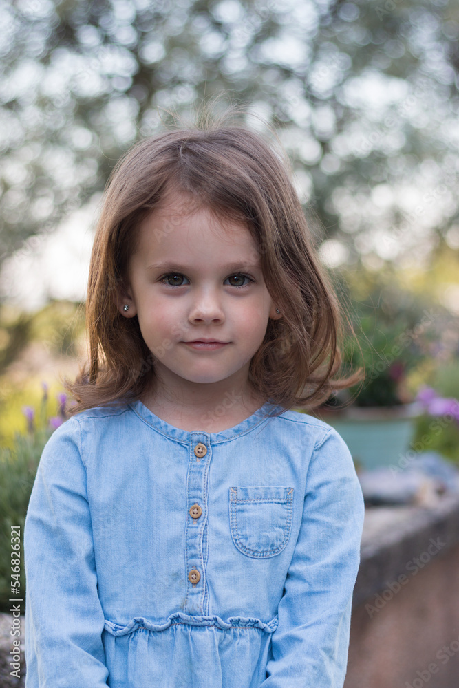 A little beautiful preschooler girl stands in a denim blue dress against the background of flowers A little beautiful preschooler girl stands in a denim blue dress against the background of flowers