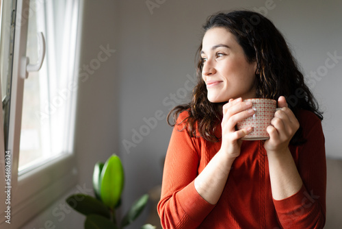 Young happy beautiful woman holding a cup of hot drink and looking trough the window