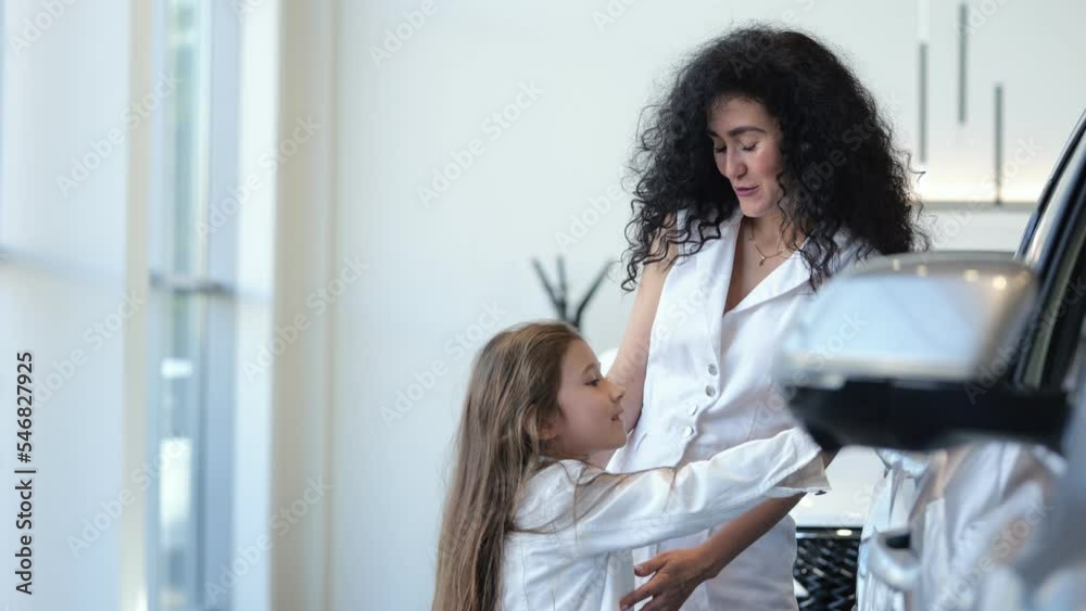 Happy mother chooses new car with excited preschooler daughter in automobile salon. Black-haired businesswoman discusses vehicle with long-haired girl