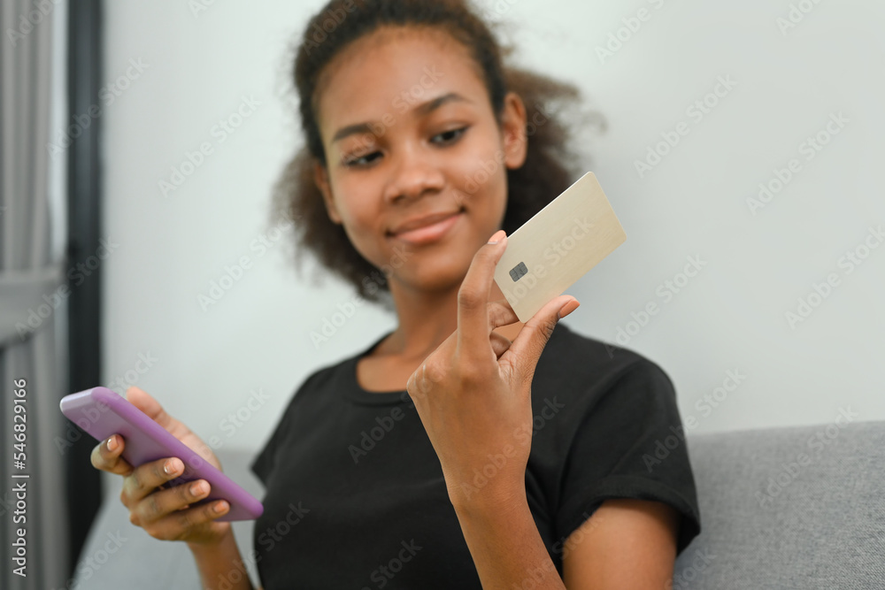 Smiling African American woman holding credit card and using smart phone. E-commerce, online shopping, internet banking concept