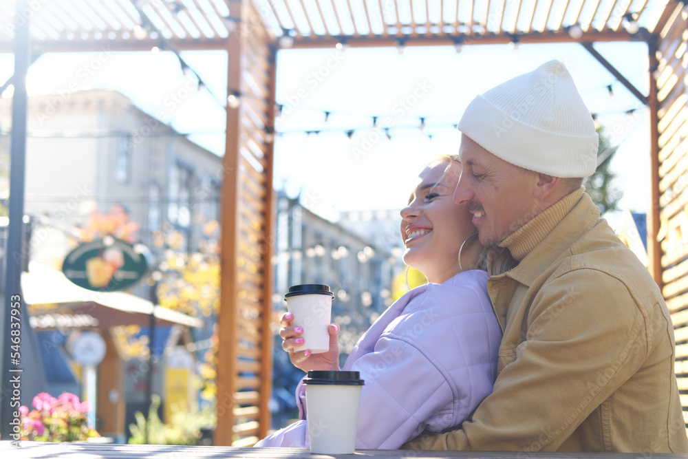 Adult guy and girl drink coffee on a street terrace in a cafe. The guy hugs the girl from behind
