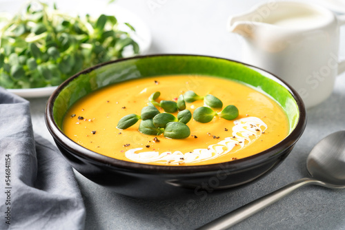 Creamy pumpkin fall soup in green bowl on the light gray table. Butternut squash cream soup garnished with heavy cream and fresh sunflower microgreen. Selective focus