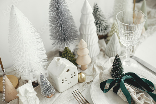 Stylish Christmas table setting. Festive napkin with ribbon and bell on plate  vintage cutlery  wineglass  modern christmas trees and houses on white rustic table. Holiday brunch