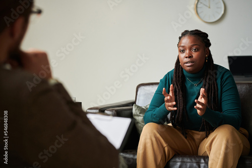 Young African American girl sitting on sofa and discussing her problems with psychologist at office photo