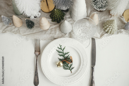 Wallpaper Mural Modern Christmas table setting. Cedar branch with bell on plate, vintage cutlery, glasses, festive little christmas trees and houses on white rustic table. Holiday arrangement of table, top view Torontodigital.ca