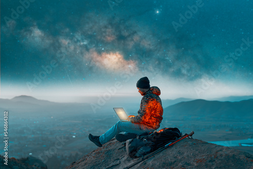 silhouette of a person sitting on top of a mountain working on his laptop at night over the valley