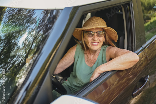 Happy senior woman driver in straw hat driving sitting in new car, smiling looking at camera enjoying journey. Driving courses and life insurance. Retired people activity and road trip concept 