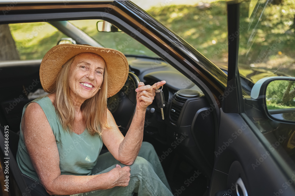 Naklejka premium Excited senior woman sitting in new car outdoors holding keys, smiling, enjoying newly bought auto. Driving courses, insurance, road trip. Retired people activity. Happy female client car dealership