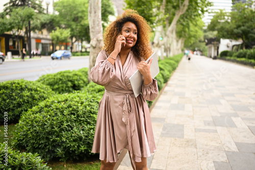 Attractive happy stylish plus size African American woman student freelancer afro hair with laptop computer outside. Young female office worker speaks phone in big urban city summer street. Diversity