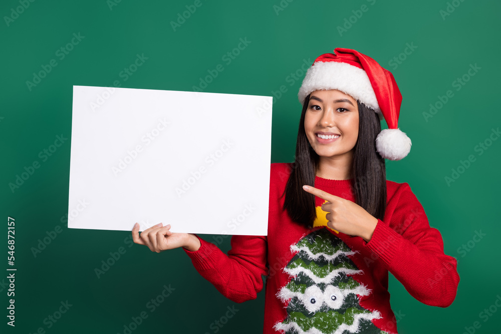 Photo of funny excited vietnamese lady wear red santa claus hat finger direct empty paper blank useful information isolated on green color background Photo of funny excited vietnamese lady wear red santa claus hat finger direct empty paper blank useful information isolated on green color background