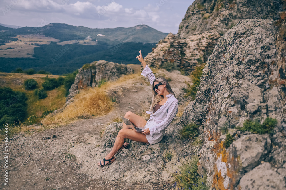 Naklejka premium Young woman freelancer traveler working online anywhere outdoors using laptop enjoying mountain peak view. Happy female downshifter in sunglasses holding computer in unusual workplace at summer