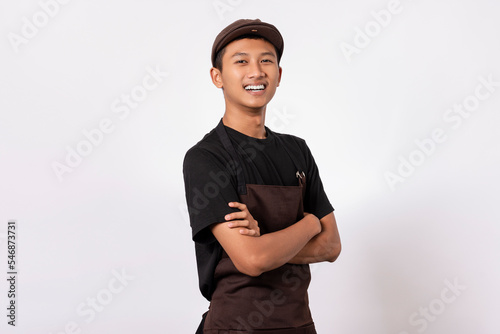 Handsome barista asian man wearing brown apron and black t-shirt isolated over white background standing with crossed arms smiling confident looking at camera