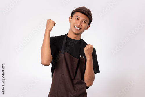 Handsome barista asian man wearing brown apron and black t-shirt isolated over white background very happy and excited celebrating winner with arms raised. success winning concept.