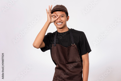 Handsome barista asian man wearing brown apron and black t-shirt isolated over white background smiling doing ok sign with hand and fingers. ok gesture.