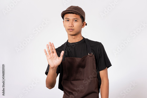 Handsome barista asian man wearing brown apron and black t-shirt isolated over white background serious expression doing stop sign with palm of the hand