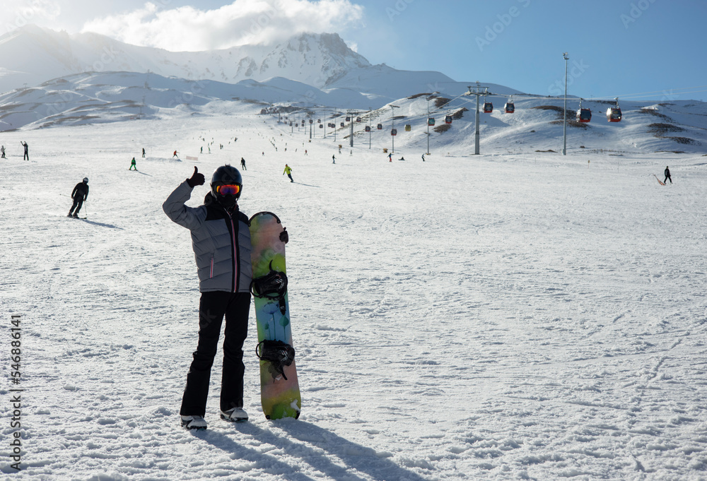 A girl with snowboard dressed in a ski suit and helmet stands against the backdrop of snow-covered mountain ski slope and a bright blue sky.  Winter. Sport and travel content  