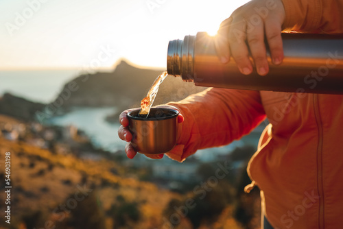 A human pours hot tea from a thermos into a mug on the mountain. Concept of autumn outdoor recreation. 