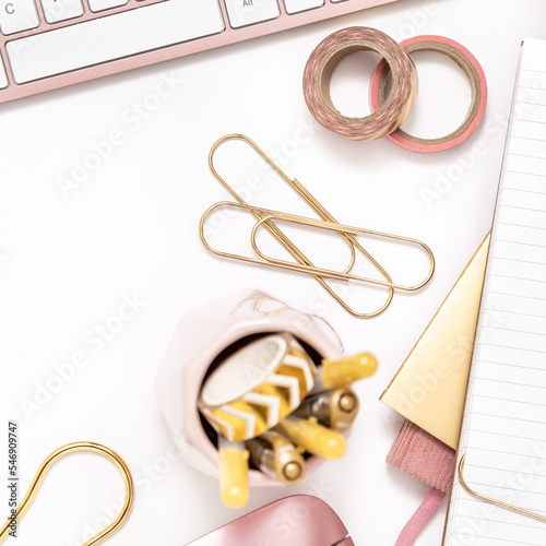 A femininely styled desktop in shades in gold and dusty pink with modem stationery. Lifestyle theme inspired by the office workspace of a stylish woman. Flat lay with fresh flowers pink keyboard and photo