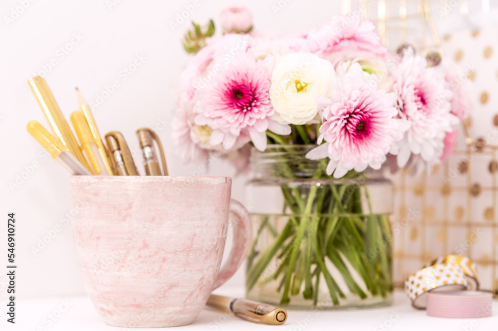 A femininely styled desktop in shades in gold and dusty pink with modem stationery. Lifestyle theme inspired by the office workspace of a stylish woman. With fresh flowers including Ranuculus