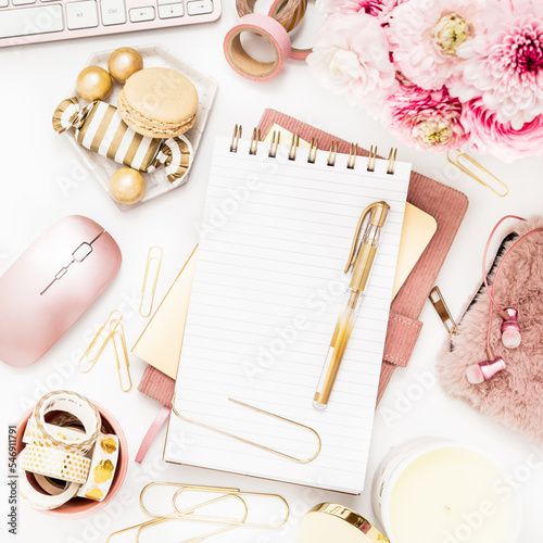 A femininely styled desktop in shades in gold and dusty pink with modem stationery. Lifestyle theme inspired by the office workspace of a stylish woman. Flat lay with fresh flowers pink keyboard and photo