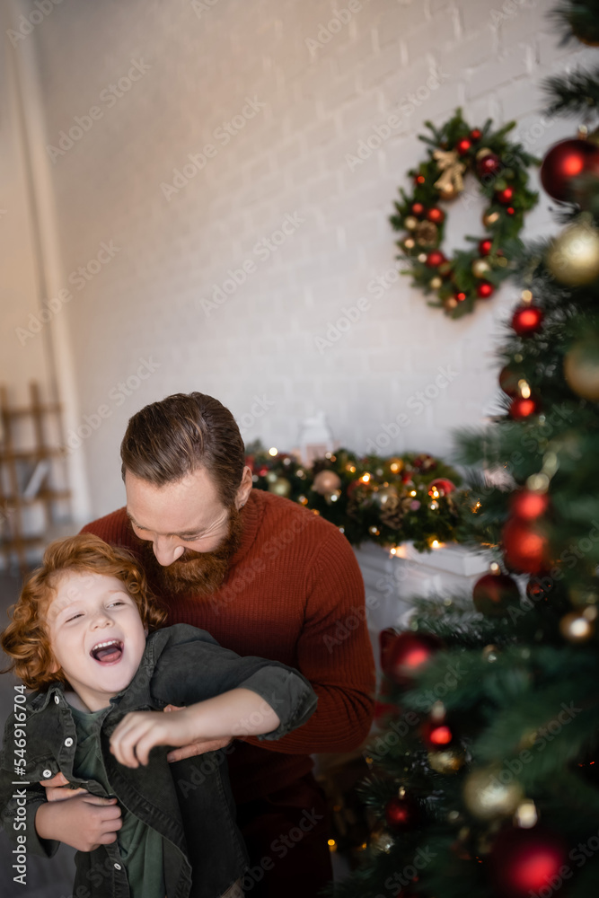 man playing with laughing redhead son near blurred Christmas decor man playing with laughing redhead son near blurred Christmas decor