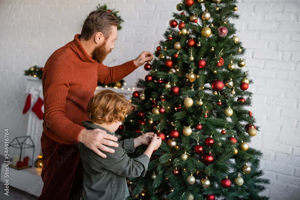 redhead kid with bearded father decorating Christmas tree in living room at home redhead kid with bearded father decorating Christmas tree in living room at home