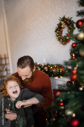 man playing with laughing redhead son near blurred Christmas decor