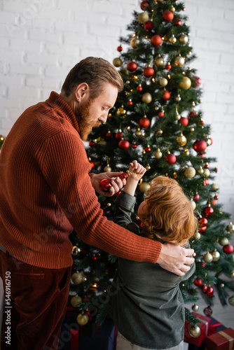 bearded man holding bauble near redhead son decorating Christmas tree