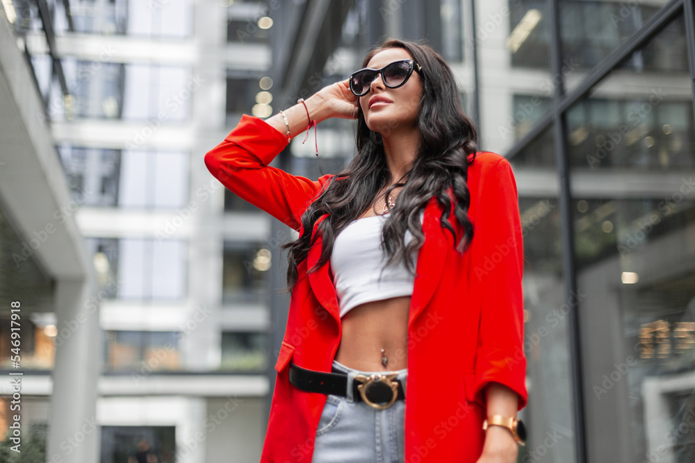 Beautiful successful fashionable business woman with curly hair and fashion sunglasses wearing a fashion red blazer, top, jeans and a purse walks in the city near modern buildings