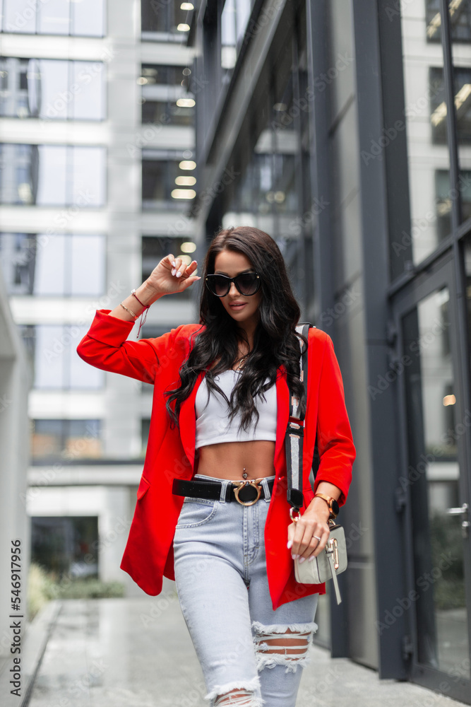 Stylish casual pretty woman in fashionable urban outfit with a red blazer, jeans and a white top with a purse walking in the city and wearing sunglasses