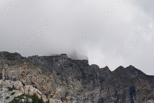 Rocky walls of mountains in low angle view in Switzerland. The upper part is covered in clouds and fog. There is some vegetation on the rocks. There is a lot of copy space im upper half of the photo. 