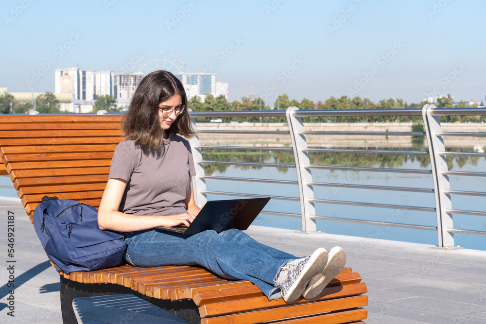 A Spanish student girl with glasses typing text on a laptop, looking for something on the Internet, chatting online with friends while sitting outdoors in the park. Freelance. Online learning concept