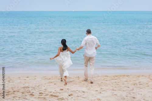 Happy wedding couple running by the beach. Romantic feelings and vibes. Back view of a romantic couple walking at beach.