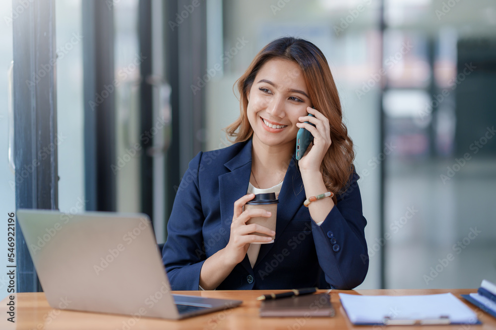 Asian businesswoman sitting with her laptop happily chatting on the phone with a customer explaining in detail about her work and company products in the office. Asian businesswoman sitting with her laptop happily chatting on the phone with a customer explaining in detail about her work and company products in the office.