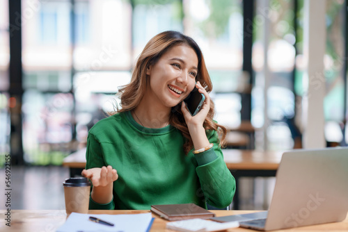Εκτύπωση καμβά Asian businesswoman sitting with her laptop happily chatting on the phone with a customer explaining in detail about her work and company products in the office