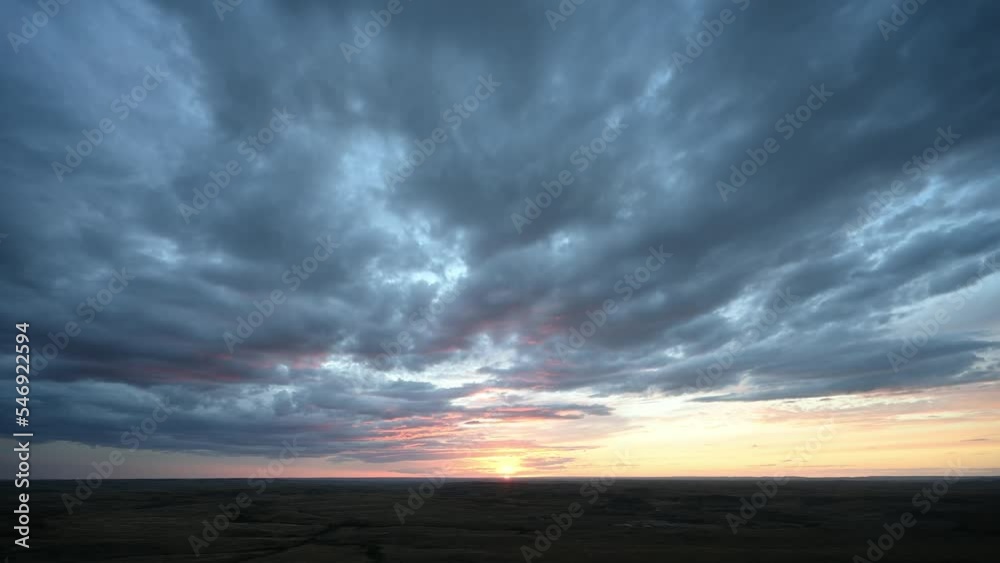 Time lapse of a sunset over prairie badlands with teal blue to orage colored clouds moving towards the viewer.
