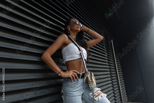 Beautiful stylish woman with a slender tanned body in fashion summer clothes with a white top and ripped jeans with a fashionable handbag near a black metal wall. Urban pretty girl with sunglasses