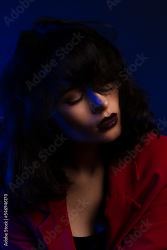 Close up portrait of girl with makeup and hairstyle  wear red suit  over blue neon colourful studio light.