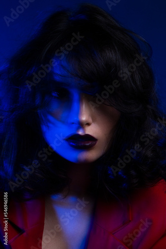 Close up portrait of girl with makeup and hairstyle, wear red suit, looking at camera, over blue neon studio light.