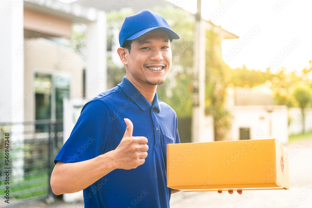Portrait of handsome delivery asian man wearing blue uniform holding cardboard boxes in front of customer house. He smiling and thumb up to camera feeling happy with his job. Portrait of handsome delivery asian man wearing blue uniform holding cardboard boxes in front of customer house. He smiling and thumb up to camera feeling happy with his job.